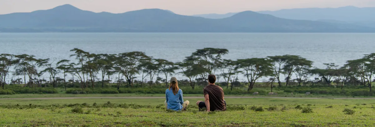 Two visitors sitting on the grass looking out across Lake Naivasha towards the hills